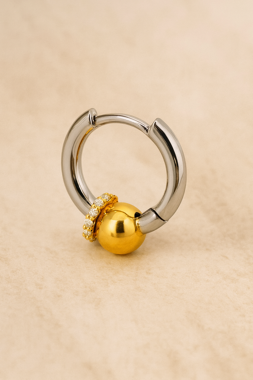 Two-tone Kora piercing bead huggie earring in sterling silver with gold bead and CZ halo on beige marble background