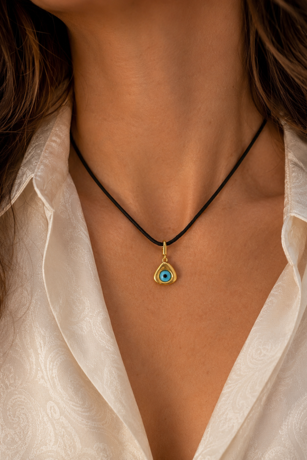 A close-up of a person wearing a black cord necklace with a gold teardrop-shaped pendant featuring a blue and white evil eye design, paired with a white collared shirt.