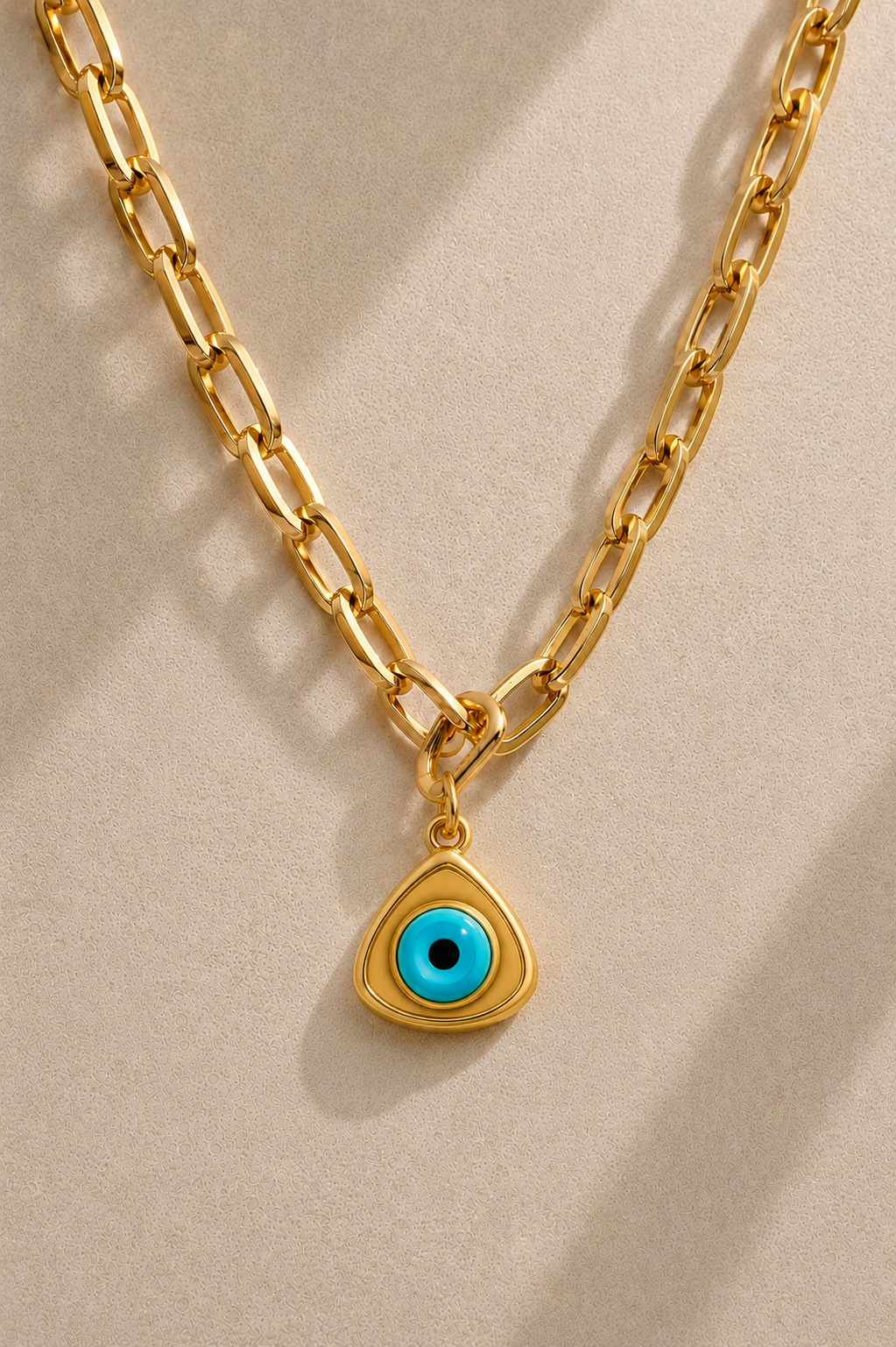 Cara chunky gold paperclip chain necklace with triangle evil eye charm on beige background