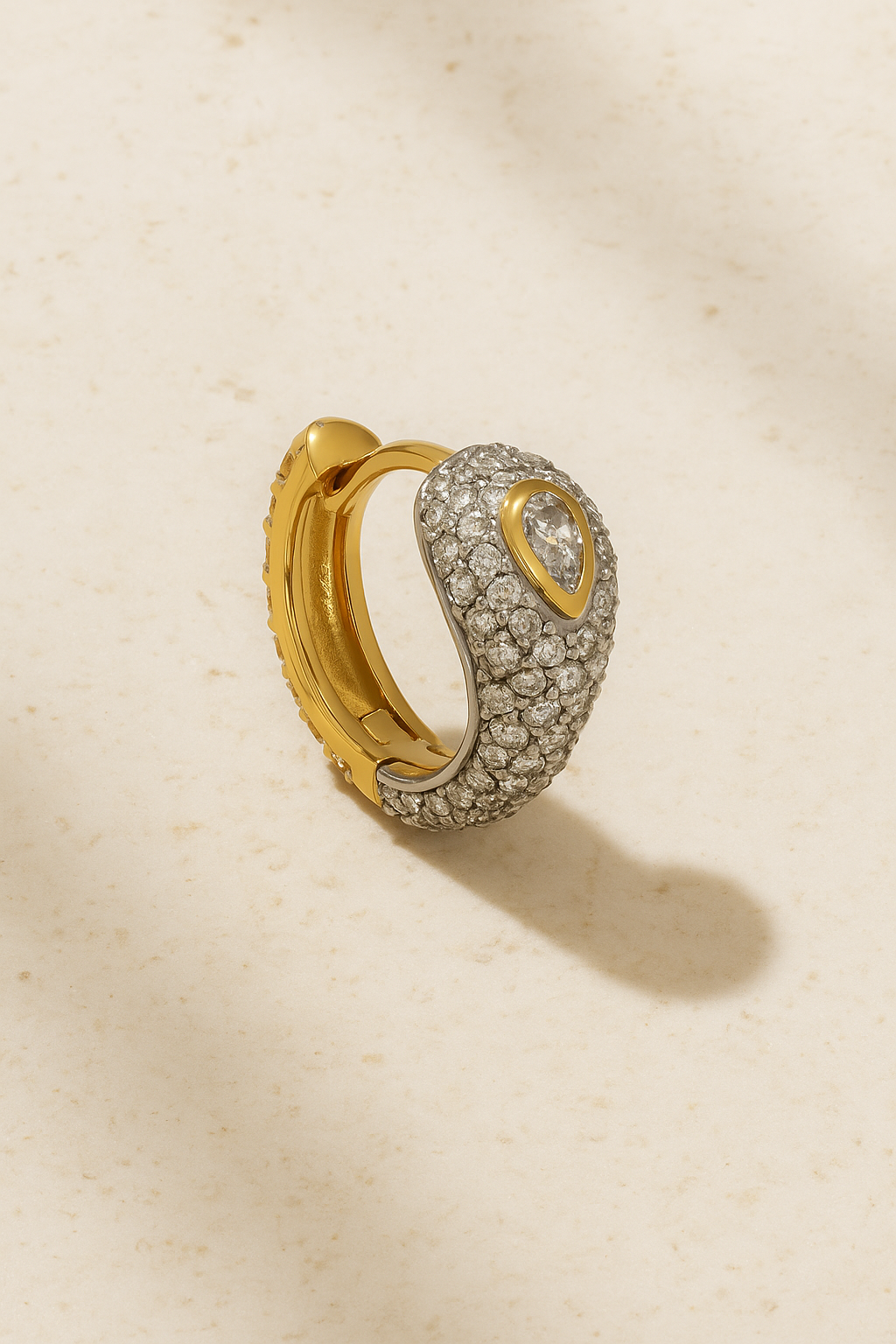 Gold Ophelia pavé huggie with pear-cut centre stone displayed on warm stone background.