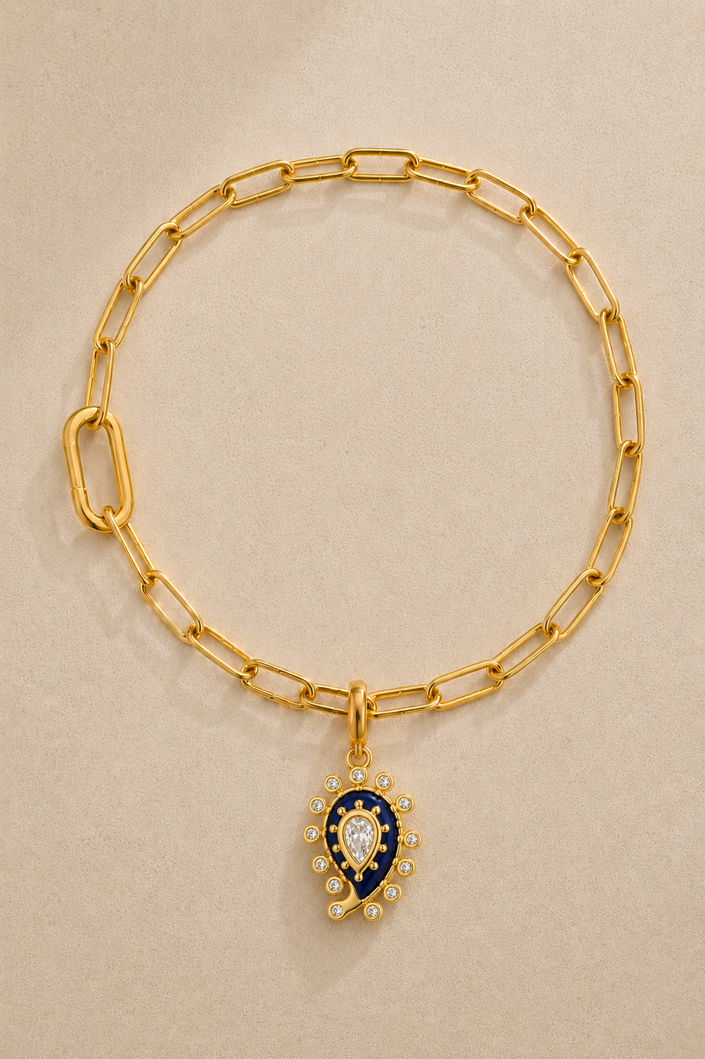 Gold paperclip bracelet with lapis paisley evil eye charm on beige background