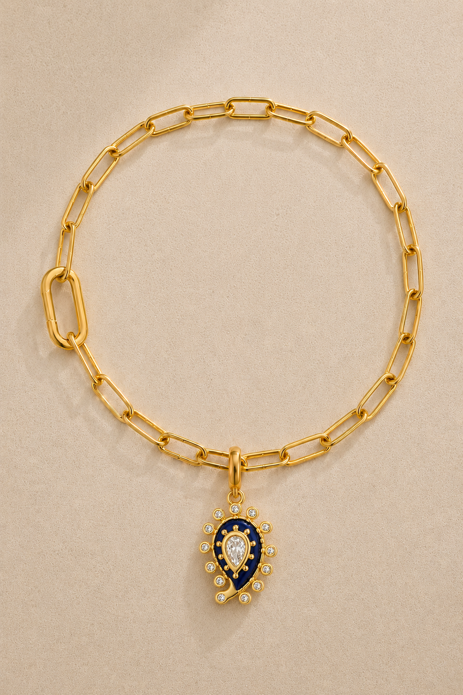 Gold paperclip bracelet with lapis paisley evil eye charm on beige background
