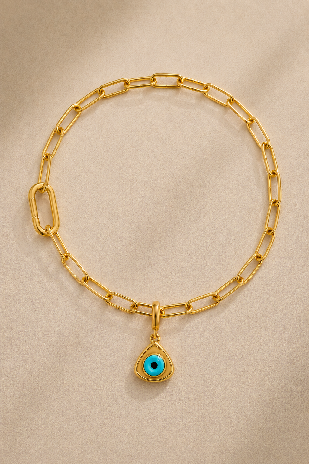 Gold paperclip bracelet with turquoise triangle evil eye charm on beige background