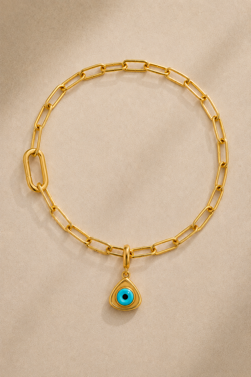 Gold paperclip bracelet with turquoise triangle evil eye charm on beige background