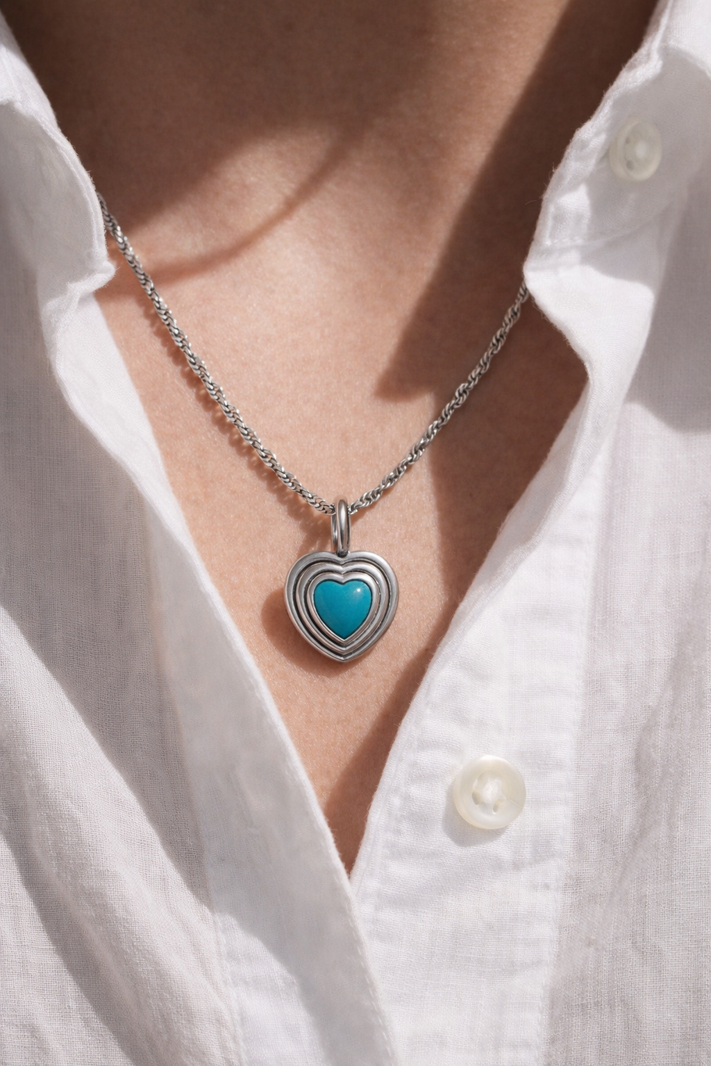 Turquoise heart pendant necklace on slim twisted sterling silver chain worn over a crisp white shirt, studio lighting