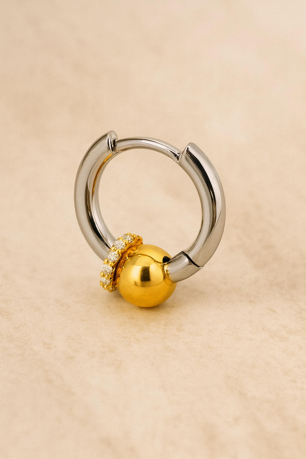 Two-tone Kora piercing bead huggie earring in sterling silver with gold bead and CZ halo on beige marble background